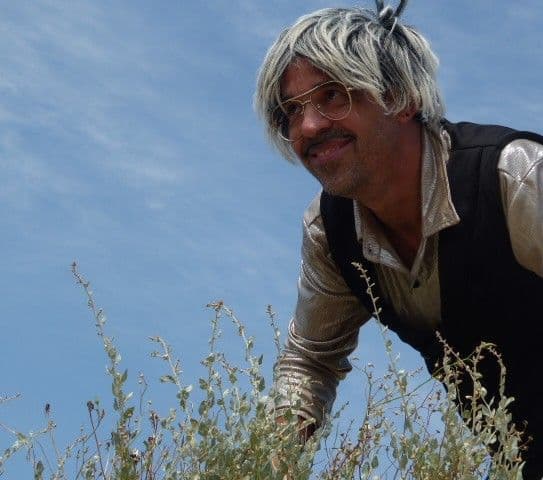 OseteDelirios filmeko aktorea. Ileordea eta betaurrekoak ditu ezaugarri, eta belar artean ageri da ingurune natural batean.